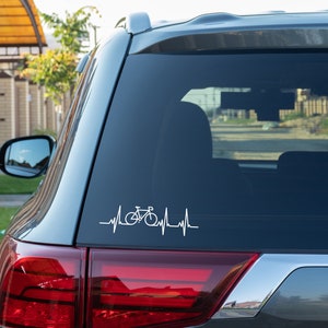 Op de afbeelding: Witte vinyl sticker van een fiets geïntegreerd in een hartslaglijn, bevestigd op de achterruit van een voertuig. Het ontwerp suggereert een passie voor fietsen. Het achterlicht van de auto is eronder zichtbaar.