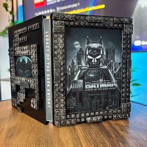 Puede incluir: Una caja de presentación negra con un gráfico de LEGO Batman y el texto LEGO BATMAN LEGACY OF THE DARK KNIGHT. El marco está construido con elementos texturizados de color gris oscuro similares a ladrillos. El panel lateral muestra el texto.
