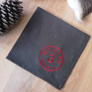 Op de afbeelding: Grijze linnen servet met een rode geborduurde cirkel met de tekst "MOUFLON 3 MERIBEL".