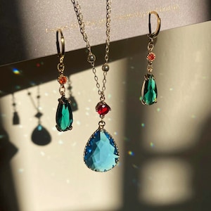 Op de afbeelding: Een goudkleurige sieradenset met een ketting en oorbellen. De ketting heeft een grote, lichtblauwe druppelvormige hanger met een rode accent. De oorbellen en de ketting hebben groene en rode accenten. De set wordt gepresenteerd tegen een neutrale achtergrond.