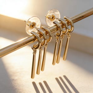 Könnte beinhalten: Zwei goldfarbene Creolen, die jeweils mit drei hängenden vertikalen Stabanhängern versehen sind. Die Ohrringe sind mit transparenten Kunststoff-Steckern gesichert und hängen an einem dünnen, horizontalen goldfarbenen Metallstab.