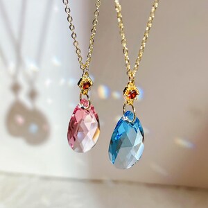 May include: Two gold necklaces with teardrop pendants. One pendant is light pink, the other is blue. Each faceted pendant hangs from a gold chain. The pendants are accented with small, red-orange gemstones.