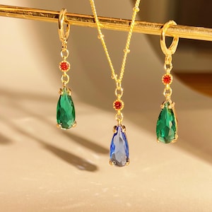 Puede incluir: Un conjunto de joyas doradas con un collar y dos pendientes. El collar tiene un colgante azul en forma de lágrima, mientras que los pendientes tienen colgantes verdes en forma de lágrima. Cada pieza tiene detalles rojos y dorados.