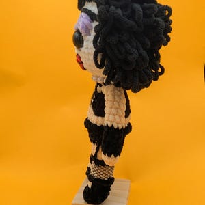 K&ouml;nnte beinhalten: Handgemachte H&auml;kelpuppe mit schwarzen Locken, einem wei&szlig;en Gesicht mit lila Lidschatten und einer roten Lippe. Die Puppe tr&auml;gt ein schwarz-wei&szlig;es Outfit, Netzstr&uuml;mpfe und schwarze Stiefel. Sie steht auf einem kleinen Holzsockel vor gelbem Hintergrund.