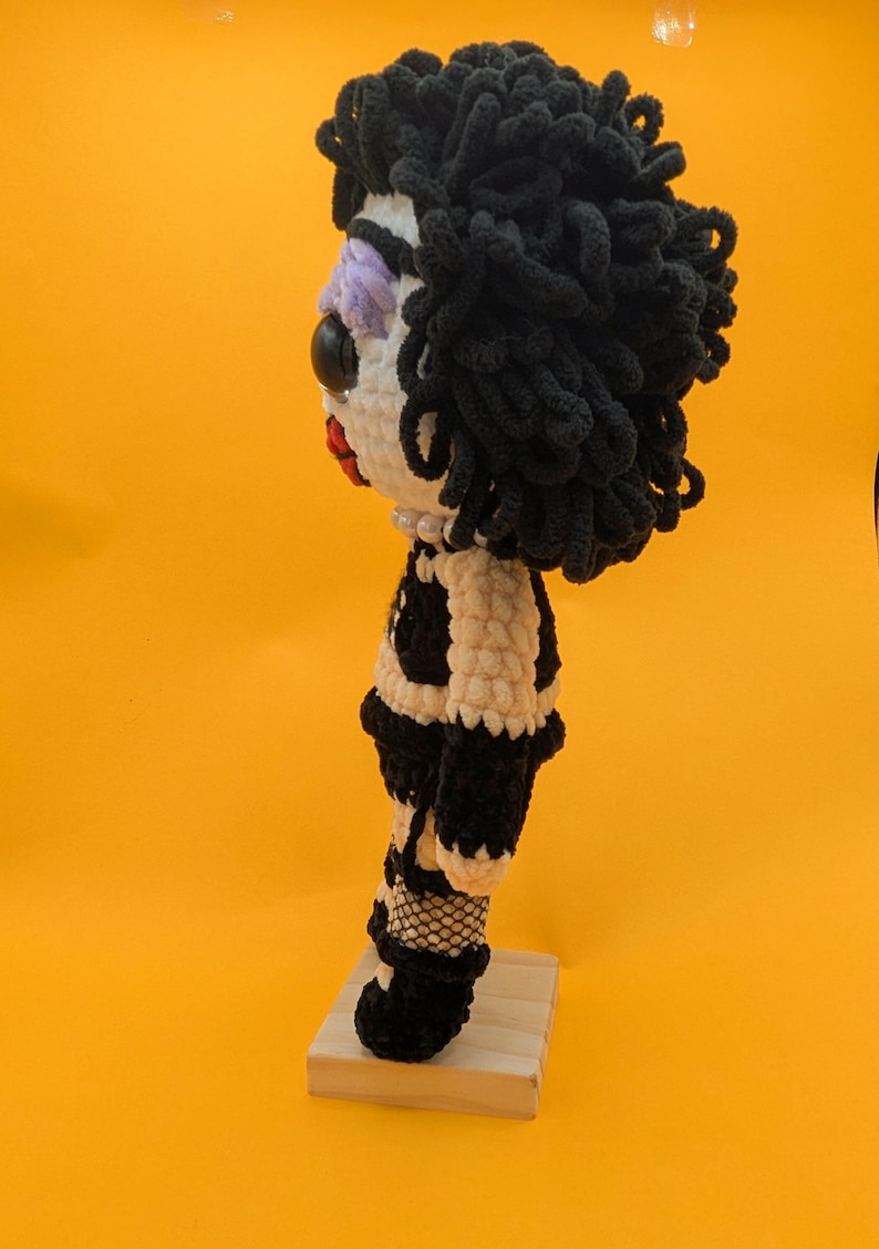 K&ouml;nnte beinhalten: Handgemachte H&auml;kelpuppe mit schwarzen Locken, einem wei&szlig;en Gesicht mit lila Lidschatten und einer roten Lippe. Die Puppe tr&auml;gt ein schwarz-wei&szlig;es Outfit, Netzstr&uuml;mpfe und schwarze Stiefel. Sie steht auf einem kleinen Holzsockel vor gelbem Hintergrund.