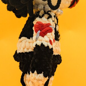 K&ouml;nnte beinhalten: Handgemachte Puppe in Schwarz und Creme. Die Puppe hat einen schwarzen Kopf und Haare, einen cremefarbenen K&ouml;rper mit schwarzen Akzenten und ein rotes herzf&ouml;rmiges Detail auf der Brust. Der Hintergrund ist gelb.