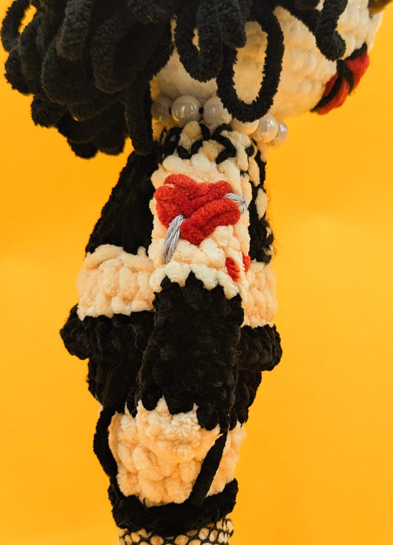 K&ouml;nnte beinhalten: Handgemachte Puppe in Schwarz und Creme. Die Puppe hat einen schwarzen Kopf und Haare, einen cremefarbenen K&ouml;rper mit schwarzen Akzenten und ein rotes herzf&ouml;rmiges Detail auf der Brust. Der Hintergrund ist gelb.