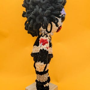 K&ouml;nnte beinhalten: Handgemachte H&auml;kelpuppe mit schwarzen Locken, einem wei&szlig;en Gesicht mit roten Lippen und einer Perlenkette. Die Puppe tr&auml;gt ein schwarz-wei&szlig;es Outfit mit rotem Herzdetail und Netzstr&uuml;mpfen. Sie steht auf einem kleinen Holzsockel vor gelbem Hintergrund.