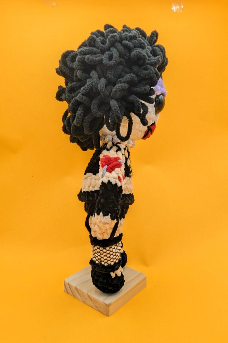 K&ouml;nnte beinhalten: Handgemachte H&auml;kelpuppe mit schwarzen Locken, einem wei&szlig;en Gesicht mit roten Lippen und einer Perlenkette. Die Puppe tr&auml;gt ein schwarz-wei&szlig;es Outfit mit rotem Herzdetail und Netzstr&uuml;mpfen. Sie steht auf einem kleinen Holzsockel vor gelbem Hintergrund.