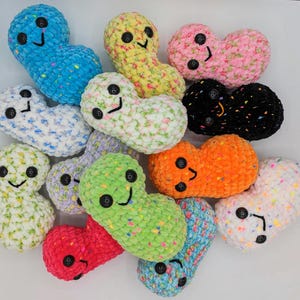 May include: A collection of 12 crocheted bean-shaped plushies with smiling faces. The plushies are in various colours, including blue, yellow, pink, white, green, orange, and black.