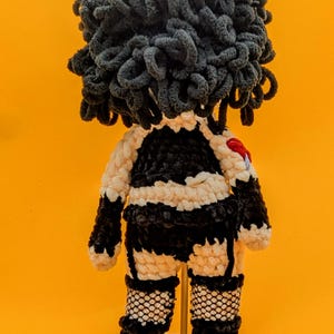 K&ouml;nnte beinhalten: Handgemachte H&auml;kelpuppe mit einem schwarz-wei&szlig;en Outfit, Netzstr&uuml;mpfen und gro&szlig;en, dunkelgrauen, lockigen Haaren. Die Puppe hat ein kleines rotes Herz auf dem R&uuml;cken. Sie wird auf einem kleinen Holzst&auml;nder vor einem leuchtend orangefarbenen Hintergrund pr&auml;sentiert.