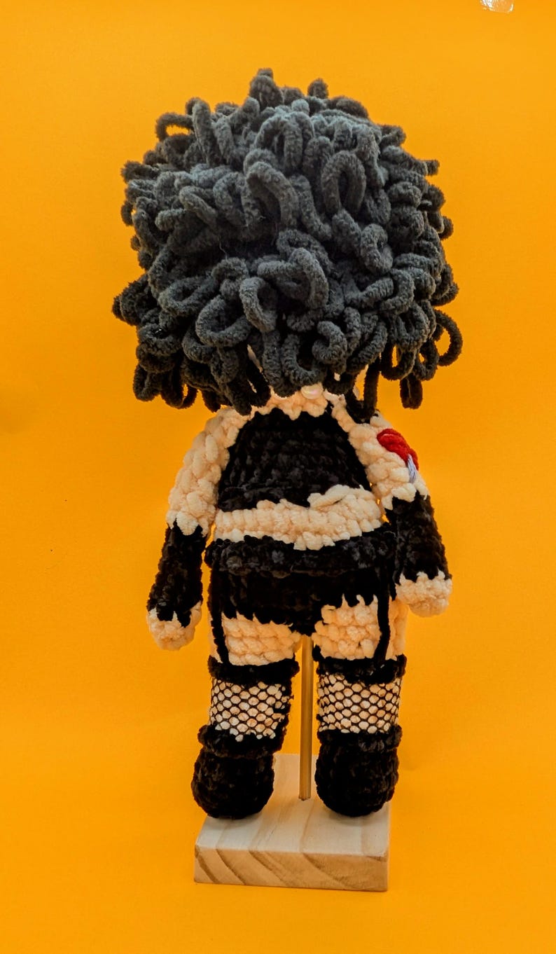 K&ouml;nnte beinhalten: Handgemachte H&auml;kelpuppe mit einem schwarz-wei&szlig;en Outfit, Netzstr&uuml;mpfen und gro&szlig;en, dunkelgrauen, lockigen Haaren. Die Puppe hat ein kleines rotes Herz auf dem R&uuml;cken. Sie wird auf einem kleinen Holzst&auml;nder vor einem leuchtend orangefarbenen Hintergrund pr&auml;sentiert.