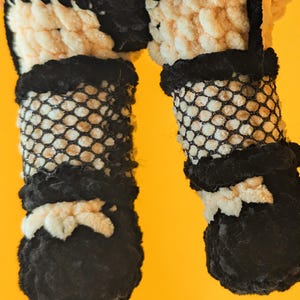 K&ouml;nnte beinhalten: Nahaufnahme der Beine und F&uuml;&szlig;e einer handgefertigten H&auml;kelpuppe. Die Beine der Puppe sind mit einem schwarzen Fischernetzmaterial bedeckt, mit schwarzen und wei&szlig;en H&auml;keldetails. Die F&uuml;&szlig;e sind schwarz mit wei&szlig;en Akzenten, vor einem gelben Hintergrund.