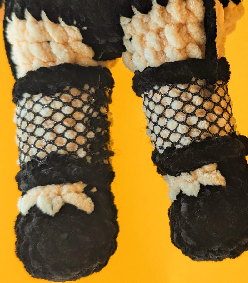 K&ouml;nnte beinhalten: Nahaufnahme der Beine und F&uuml;&szlig;e einer handgefertigten H&auml;kelpuppe. Die Beine der Puppe sind mit einem schwarzen Fischernetzmaterial bedeckt, mit schwarzen und wei&szlig;en H&auml;keldetails. Die F&uuml;&szlig;e sind schwarz mit wei&szlig;en Akzenten, vor einem gelben Hintergrund.