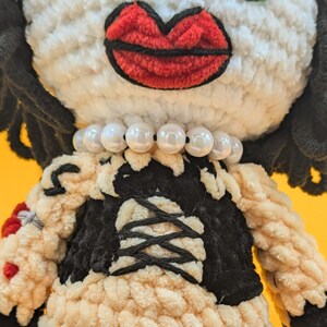 K&ouml;nnte beinhalten: Geh&auml;kelte Puppe mit wei&szlig;em Gesicht, gro&szlig;en roten Lippen und schwarzen Haaren. Die Puppe tr&auml;gt ein schwarz-cremefarbenes Kleid mit einer Perlenkette. Die Augen der Puppe sind gro&szlig; und gl&auml;nzend.