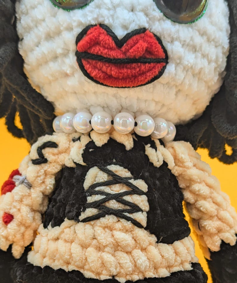 K&ouml;nnte beinhalten: Geh&auml;kelte Puppe mit wei&szlig;em Gesicht, gro&szlig;en roten Lippen und schwarzen Haaren. Die Puppe tr&auml;gt ein schwarz-cremefarbenes Kleid mit einer Perlenkette. Die Augen der Puppe sind gro&szlig; und gl&auml;nzend.