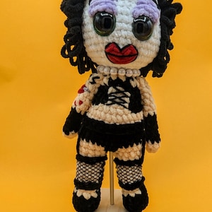 K&ouml;nnte beinhalten: Geh&auml;kelte Puppe mit wei&szlig;em Gesicht, schwarzen Locken und gro&szlig;en schwarzen Augen. Die Puppe hat rote Lippen, lila Lidschatten und eine Perlenkette. Sie tr&auml;gt ein schwarz-wei&szlig; gestreiftes Oberteil, schwarze Netzstr&uuml;mpfe und schwarze Schuhe.