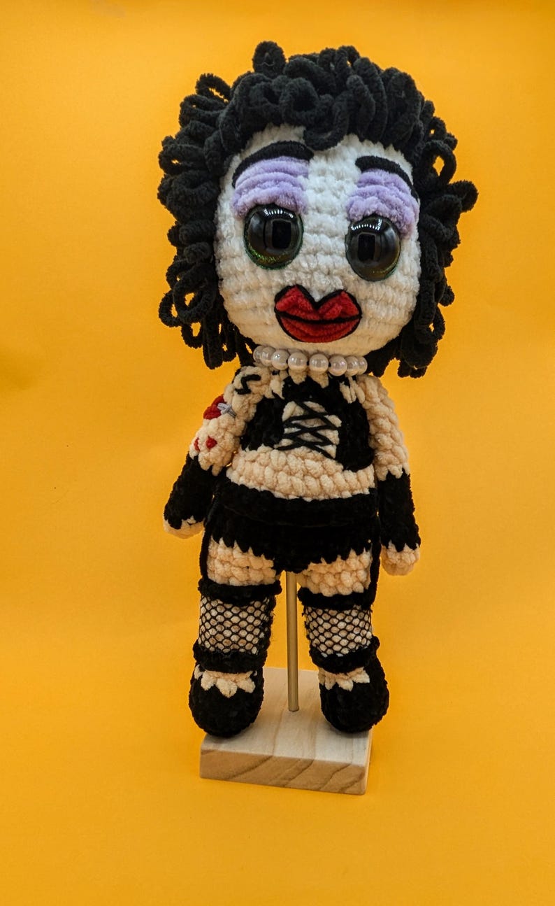 K&ouml;nnte beinhalten: Geh&auml;kelte Puppe mit wei&szlig;em Gesicht, schwarzen Locken und gro&szlig;en schwarzen Augen. Die Puppe hat rote Lippen, lila Lidschatten und eine Perlenkette. Sie tr&auml;gt ein schwarz-wei&szlig; gestreiftes Oberteil, schwarze Netzstr&uuml;mpfe und schwarze Schuhe.