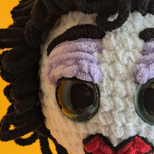 K&ouml;nnte beinhalten: Eine handgefertigte Pl&uuml;schpuppe mit schwarzen Garnhaaren, lavendelfarbenem Lidschatten und roten Lippen. Die Puppe hat gro&szlig;e, reflektierende Augen mit einem glitzernden gr&uuml;nen Rand. Das Gesicht der Puppe ist wei&szlig; und sie hat eine Perlenkette.