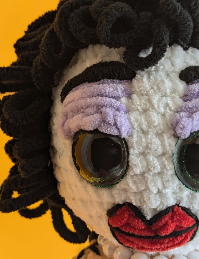 K&ouml;nnte beinhalten: Eine handgefertigte Pl&uuml;schpuppe mit schwarzen Garnhaaren, lavendelfarbenem Lidschatten und roten Lippen. Die Puppe hat gro&szlig;e, reflektierende Augen mit einem glitzernden gr&uuml;nen Rand. Das Gesicht der Puppe ist wei&szlig; und sie hat eine Perlenkette.