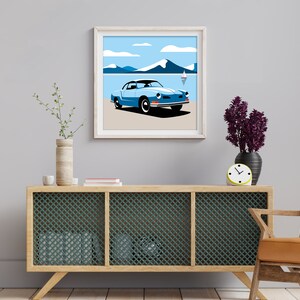 May include: A framed illustration of a blue vintage car driving on a beach with mountains in the background. The car is a Volkswagen Karmann Ghia. The illustration is in a minimalist style with flat colors and simple lines.