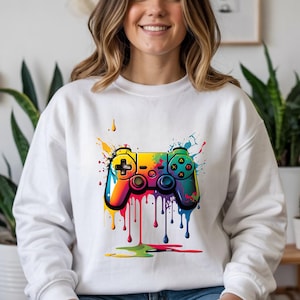 Pode incluir: Camisola branca com um gráfico abstrato e colorido de um controle de videogame. O design do controle usa uma paleta vibrante de arco-íris com efeitos de gotejamento de tinta. A camisola é um estilo casual e confortável.