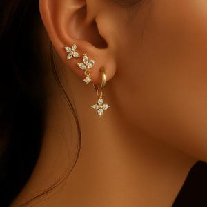Peut inclure: Boucles d'oreilles dorées avec un motif floral, ornées de pierres précieuses transparentes. L'ensemble comprend des clous d'oreilles, des boucles pendantes et des créoles. La marque "Rêve de Quartz" est visible en texte doré.