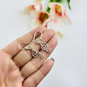 May include: Silver-coloured earrings in the shape of musical notes. The earrings are designed as treble clefs, each with a small ball at the top. The earrings are shown on a hand, with a blurred floral background.