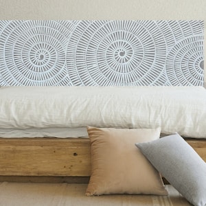 Puede incluir: Una cabecera de cama de madera con un patrón geométrico blanco de círculos concéntricos. La cabecera está unida a una cama con un edredón de lino blanco y dos almohadas decorativas.