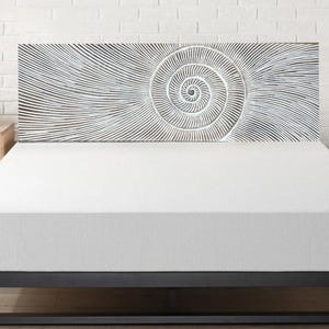 Puede incluir: Un colchón blanco sobre una estructura metálica negra, con un cabecero decorativo con un diseño en espiral. El cabecero es de madera y tiene un acabado blanco. Una pequeña mesita de noche de madera con una planta y un reloj es visible.