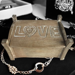 Pu&ograve; includere: Una scatola di legno con la parola "LOVE" intagliata sul coperchio. La scatola &egrave; appoggiata su una superficie nera con catene e gioielli dorati.