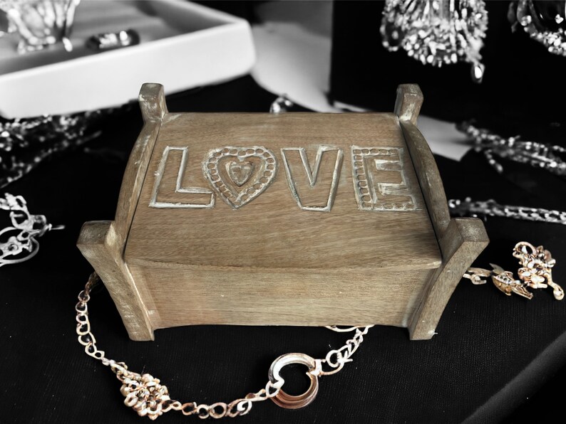 Pu&ograve; includere: Una scatola di legno con la parola "LOVE" intagliata sul coperchio. La scatola &egrave; appoggiata su una superficie nera con catene e gioielli dorati.