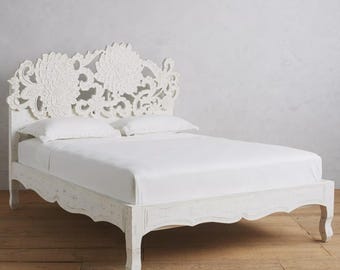 Könnte beinhalten: Weißes Holzbettgestell mit einem geschnitzten floralen Kopfteil. Das Bett hat eine Used-Optik, ein niedriges Profil und ein wellenförmiges Fußteil. Das Bett ist mit weißen Laken und Kissen bezogen.