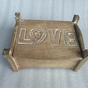 Pu&ograve; includere: Una scatola di legno con un cuore intagliato e la parola "LOVE" sul davanti. La scatola &egrave; di colore marrone chiaro e ha una finitura usurata.