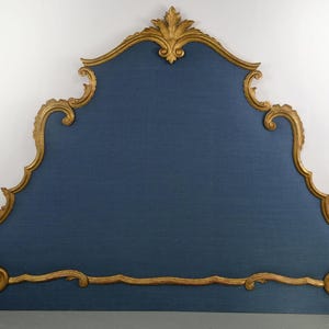 May include: A blue upholstered headboard featuring a prominent gold-colored wooden frame with intricate carved scrollwork and leaf-like detailing along the edges. The central panel is covered in a solid blue fabric with a visible woven texture.