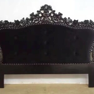 May include: Black upholstered headboard with ornate, carved wooden frame. The headboard features a curved top with decorative carvings and is accented with a row of small, silver-colored nailhead trim. The headboard is supported by two rectangular legs.