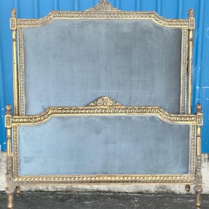 May include: An antique bed frame with a gold-colored, ornate wooden frame and a blue-gray upholstered headboard and footboard. The frame features detailed carvings and decorative accents, showcasing a classic design.