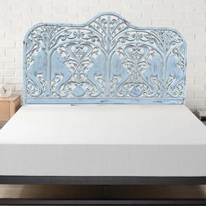 May include: A light blue headboard featuring intricate, hand-carved floral and scrollwork patterns. The wooden piece has a distressed finish, revealing natural wood tones beneath the blue paint, and is mounted behind a plain white mattress.