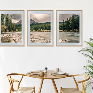 May include: Three framed landscape photographs of a river flowing through a forest with mountains in the background. The photos are hung on a white wall above a wooden table with two chairs.