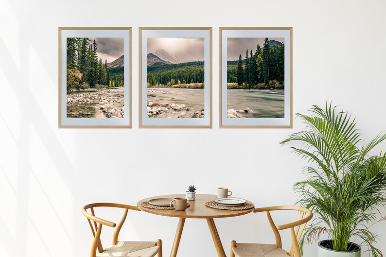 May include: Three framed landscape photographs of a river flowing through a forest with mountains in the background. The photos are hung on a white wall above a wooden table with two chairs.