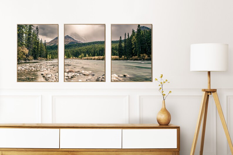 May include: Three framed landscape photographs of a river flowing through a forest with mountains in the background. The photos are hung on a white wall above a wooden dresser.