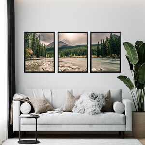May include: Three framed landscape photographs of a river flowing through a forest with mountains in the background. The photos are hung on a white wall above a white couch with pillows.