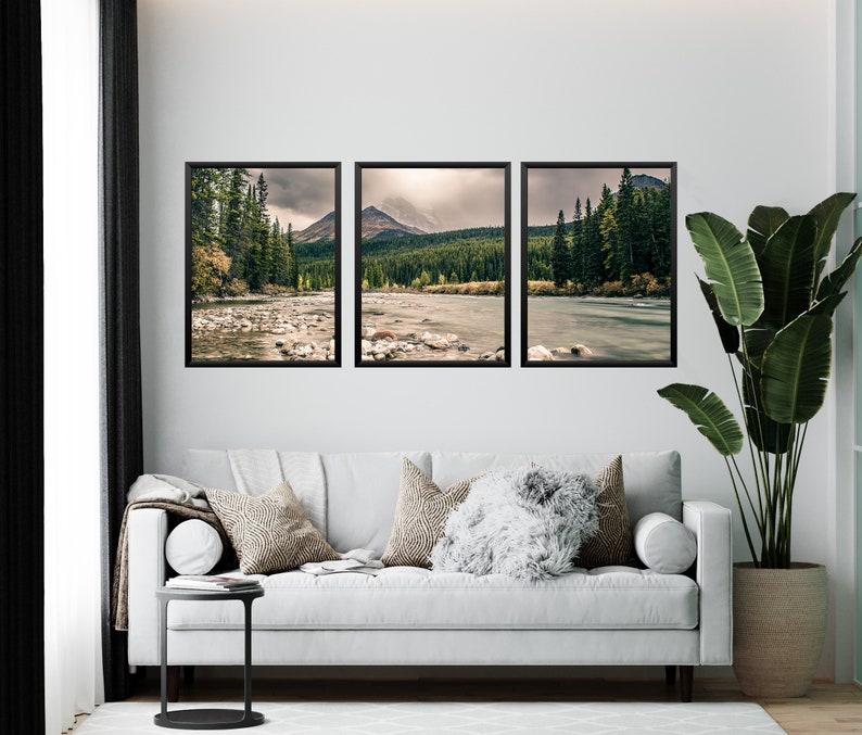 May include: Three framed landscape photographs of a river flowing through a forest with mountains in the background. The photos are hung on a white wall above a white couch with pillows.