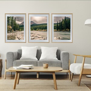 May include: Three framed landscape photographs of a river flowing through a forest with mountains in the background. The photos are hung on a white wall above a gray couch and a white chair.