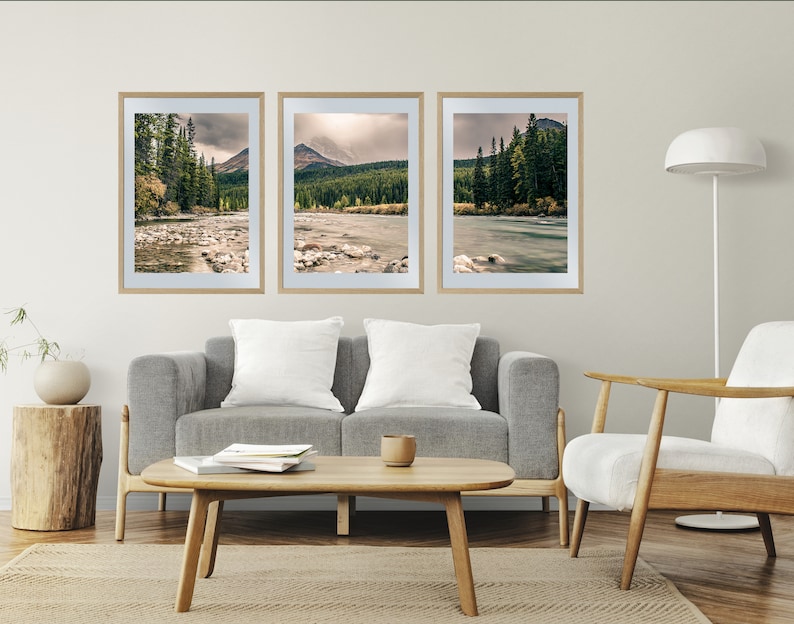 May include: Three framed landscape photographs of a river flowing through a forest with mountains in the background. The photos are hung on a white wall above a gray couch and a white chair.