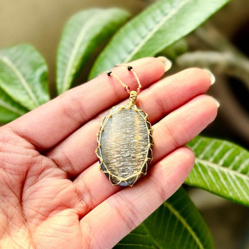 May include: An oval pendant with a dark grey and gold gemstone, wrapped in gold wire. The pendant is hanging from a cord with small beads.