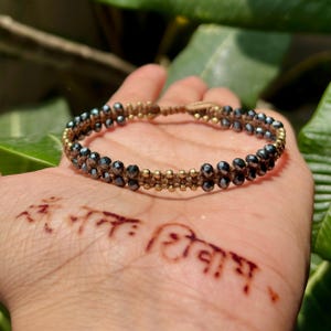 Könnte beinhalten: Ein Perlenarmband mit dunkelgrauen Perlen und goldfarbenen Akzenten. Das Armband hat ein gewebtes Design und wird auf einer Hand präsentiert. Das Armband ist ein Schmuckstück.