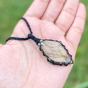 Pode incluir: Um colar de cord&atilde;o preto com um pingente de labradorita em forma de diamante envolto em cord&atilde;o preto.