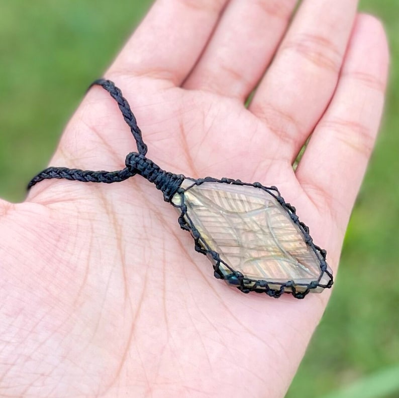 Pode incluir: Um colar de cord&atilde;o preto com um pingente de labradorita em forma de diamante envolto em cord&atilde;o preto.