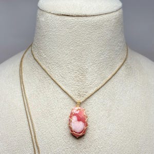 May include: A pink and white gemstone pendant necklace. The pendant is an oval shape, wrapped in gold-colored wire, and suspended from a delicate gold-colored chain. The gemstone has a marbled appearance with shades of pink and white.